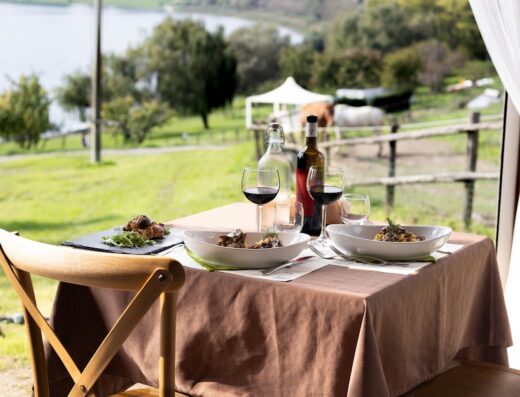 Tavolo apparecchiato con vista sul Casale di Martignano, con piatti di pasta, vino rosso e sedie di legno, per un'esperienza culinaria immersa nella natura.