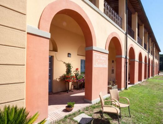 Casale La Sterpaia Hotel: vista esterna dell'hotel con archi rossi e arancioni, sedie in vimini e prato verde, ideale per un viaggio rilassante in Toscana.