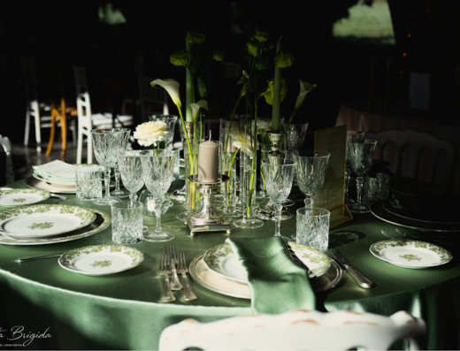 Tavolo imbandito con tovaglia verde, piatti decorati, bicchieri di cristallo, fiori bianchi e candele, ambientazione Casali Santa Brigida.