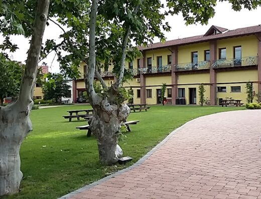 Cascina Clarabella vista esterna con prato verde, alberi e sentiero in mattoni, tavoli da picnic e edificio giallo a due piani.