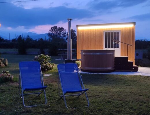 Cascina la Demilana: Vasca idromassaggio in legno accanto a una casetta di legno illuminata, con due sedie a sdraio blu in primo piano, per un relax all'aperto