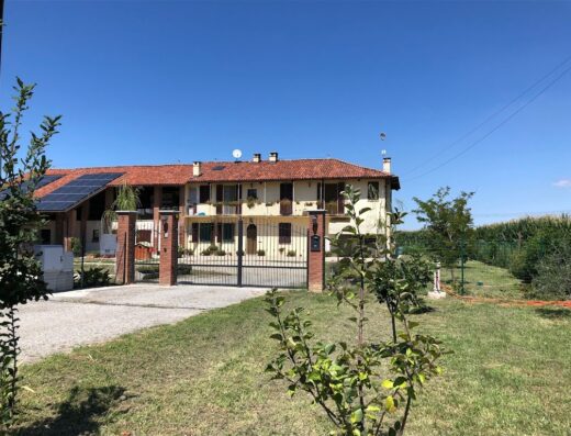 Cascina La Giardina: Facciata esterna della cascina con cancello in ferro battuto, tetto in tegole rosse, pannelli solari e giardino verde.