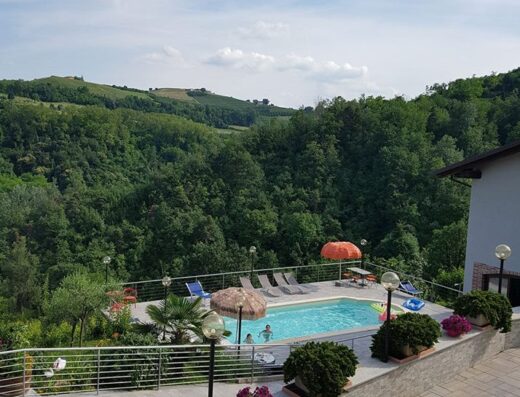 Cascina Roreto: vista panoramica sulla piscina con persone che nuotano, circondata da colline verdi e cielo azzurro. Ideale per relax e vacanze.