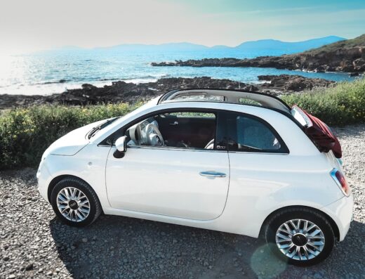 Fiat 500 cabrio bianca parcheggiata vicino alla costa rocciosa in Sardegna, ideale per escursioni e surftrip con CasualVan noleggio camper e auto.