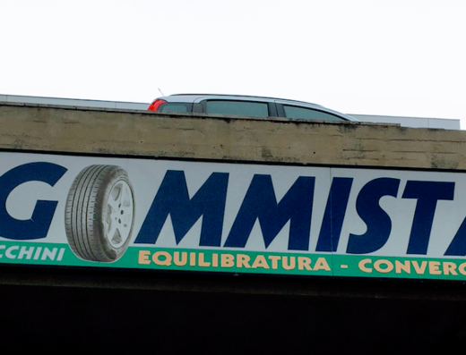 Insegna di Cecchini Gommista con pneumatico, servizi di equilibratura e convergenza per auto e moto. Sul tetto si intravede un'auto.