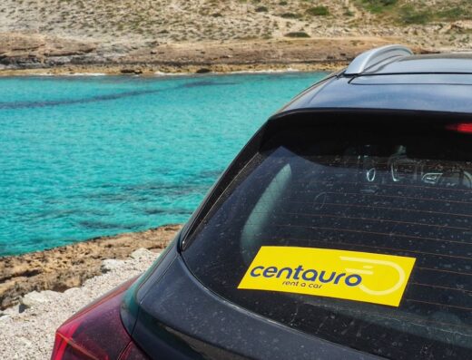 Auto Centauro Nolo con adesivo sul lunotto posteriore, sullo sfondo mare cristallino e costa rocciosa. Servizi di noleggio auto per viaggi e vacanze.
