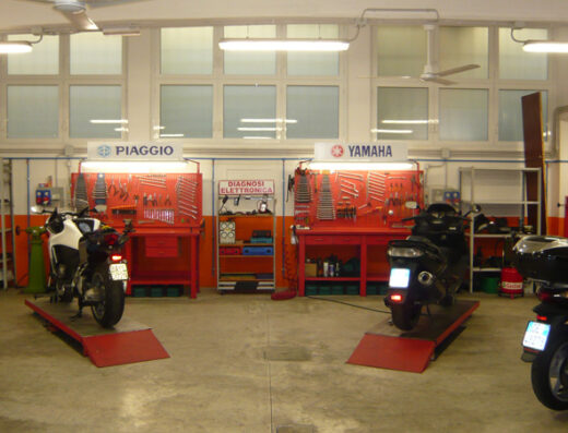 Officina moto Balduina: interni con moto Piaggio e Yamaha su piattaforme di sollevamento, banchi da lavoro con utensili per riparazione e manutenzione moto.