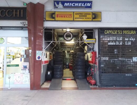 Foto del Centro Cassia Gomme, un'officina specializzata in pneumatici con marchi come Michelin e Pirelli, che offre assistenza tecnica per moto e scooter. Si vedono pile di gomme e rampe di accesso.