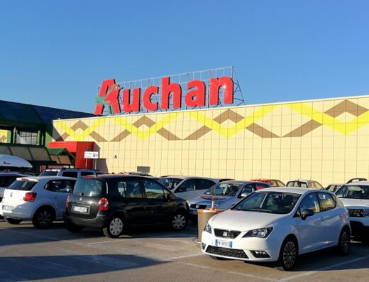 Parcheggio affollato di fronte al Centro Commerciale Emisfero Auchan Cattane, con diverse auto visibili e l'insegna del supermercato in evidenza.