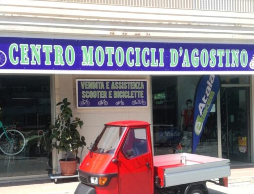 Centro Motocicli D'Agostino: un'Ape rossa parcheggiata di fronte al negozio di vendita e assistenza scooter e biciclette.