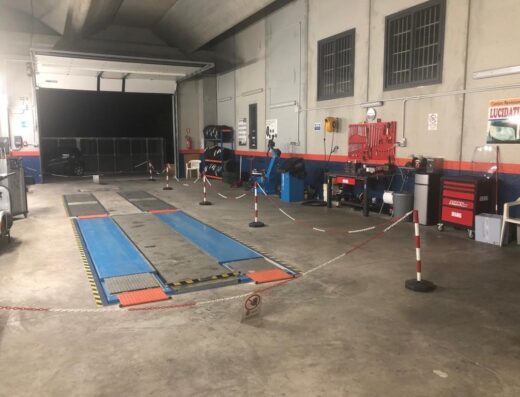 Interno di un centro revisioni auto con ponte sollevatore blu e grigio, pneumatici impilati, attrezzi e cartello "Lucidatura".