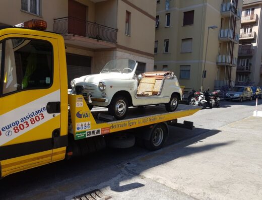 Immagine di un carro attrezzi di Centro Revisioni Ligure Srl che trasporta una Fiat 600 spiaggina bianca, auto d'epoca scoperta.