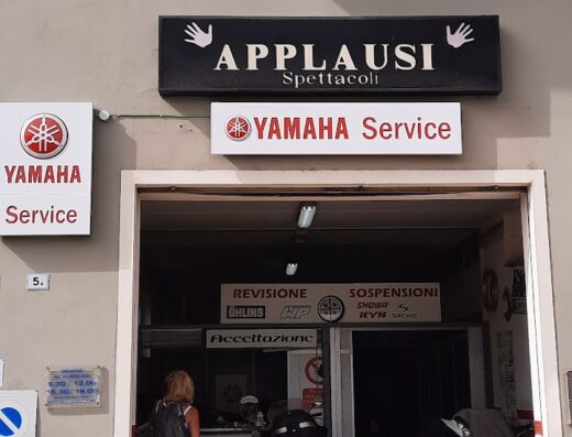 Centro Riparazione Moto Masala Roberto: facciata dell'officina Yamaha Service con moto parcheggiate, cartelli "Revisione", "Sospensioni", e donna in entrata. Servizi di riparazione e manutenzione moto.
