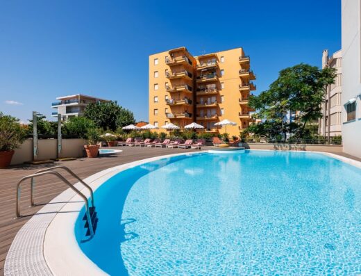 Piscina all'aperto con acqua cristallina a Pesaro, circondata da lettini prendisole e ombrelloni, con un edificio colorato sullo sfondo in una giornata di sole.