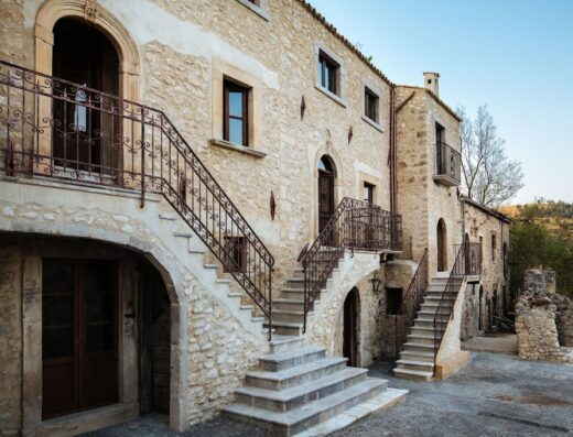 Charme in Perillis: Esterno di un edificio storico in pietra con scale esterne in ferro battuto, che conduce a diversi ingressi. L'architettura rustica si fonde con il paesaggio circostante.