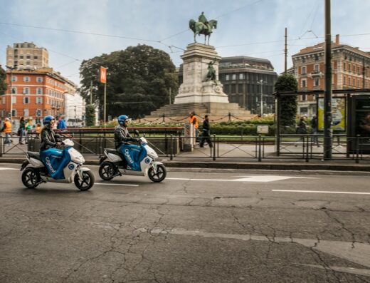 Due persone guidano scooter elettrici Cityscoot a Milano, con la statua di Garibaldi sullo sfondo. Noleggio scooter elettrico.