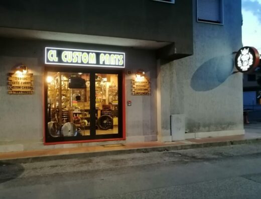 Foto della facciata di CL CUSTOM PARTS SRLS, negozio di accessori e ricambi per moto, con vetrina illuminata e insegne luminose.