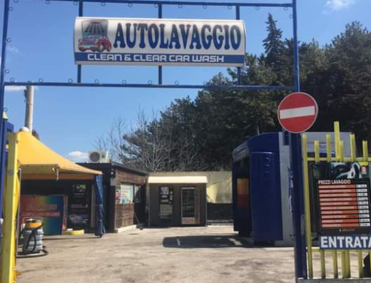 Entrance to Clean & Clear Car Wash showing the sign with prices and an 'Entrata' (Entrance) sign, with a blue and yellow gate and buildings in the background.