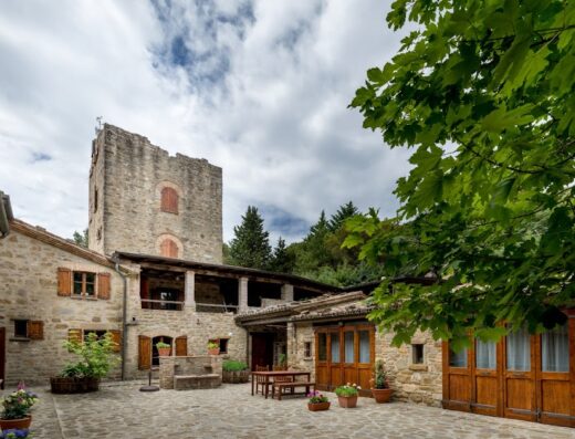 Veduta esterna di Colle Surya, una struttura in pietra con torre medievale, tavolo e sedie in cortile, vegetazione rigogliosa, cielo nuvoloso. Ideale per viaggi in moto e soggiorni unici.