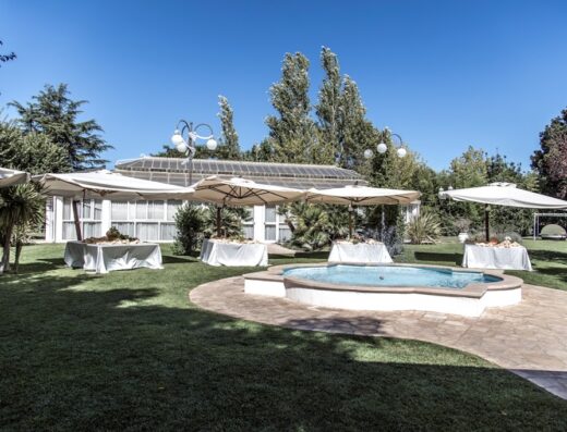 Un bellissimo giardino al Complesso Turistico Antica Masseria Dimora Mazzaro con prato verde, ombrelloni bianchi, tavoli imbanditi e una fontana centrale, ideale per eventi e relax.