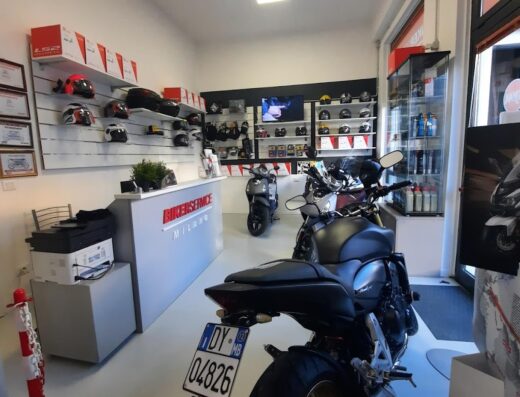 Interno del concessionario Sym Bikerservice a Milano, con moto in primo piano, caschi e accessori esposti per la vendita.