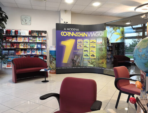 Interno dell'agenzia Cornacchini Viaggi a Modena, con divano rosso, scaffale di brochure di viaggi, cartellone pubblicitario con logo e offerte di noleggio auto e altri servizi turistici.