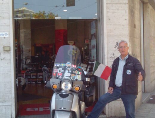 Un uomo in posa accanto a una moto d'epoca di fronte a Costantino Moto, concessionario Aprilia e Moto Guzzi, con bandiera italiana.