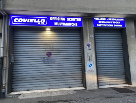 La saracinesca chiusa di Coviello Vincenzo, officina scooter multimarche dal 1978, con cartelli di tagliandi, precollaudi, restauri d'epoca e sostituzione gomme.