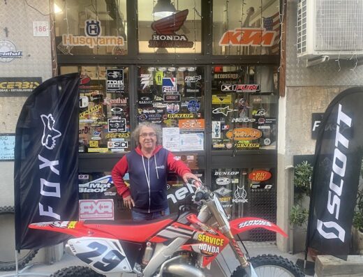 Un uomo posa con una moto Honda da cross rossa e bianca davanti al negozio Cross Action a Genova, con espositori di marchi come Husqvarna e KTM, e accessori moto come Fox e Scott visibili.