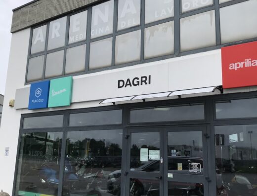 Concessionaria Dagri con vetrine che espongono moto Piaggio, Vespa, Aprilia e Moto Guzzi. Arena Medicina del Lavoro visibile in alto.
