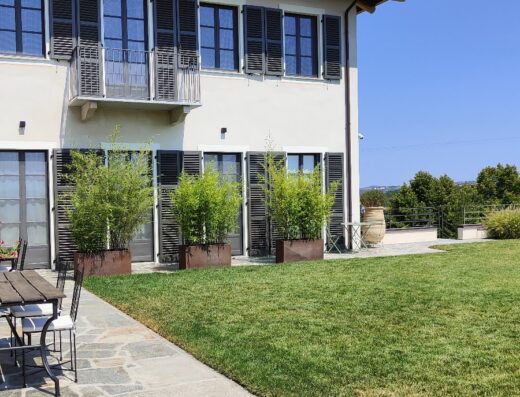 Casa di campagna con giardino curato e pavimentazione in pietra, con tavoli e sedie in ferro battuto. La facciata presenta finestre con persiane scure e balcone.