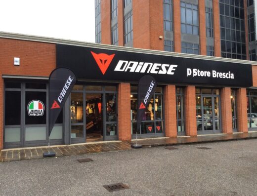 Foto esterna del Dainese D Store Brescia, negozio di abbigliamento e accessori moto. Insegna Dainese ben visibile e bandiere AGU sulla facciata.