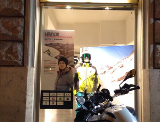Ingresso del negozio Dainese Roma con moto parcheggiata davanti. Vetrina con poster evento invernale e abbigliamento moto e sci esposto.