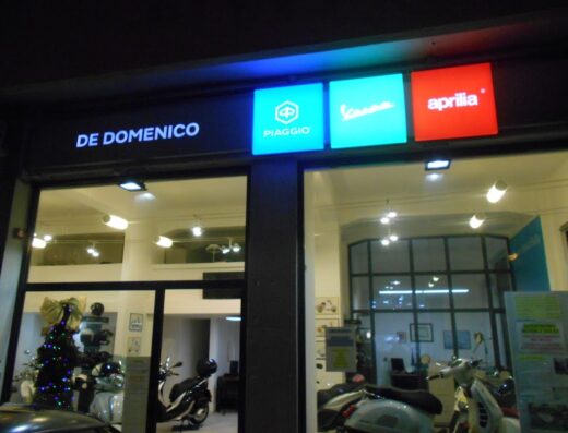 Concessionaria De Domenico SRL con insegne luminose di Piaggio, Vespa e Aprilia. Vetrina con scooter e moto in esposizione.
