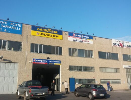De Luca Gomme: Esterno dell'officina con insegne Goodyear, Dunlop e Super Service, auto parcheggiate e personale visibile, servizio pneumatici e accessori moto