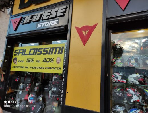 Vetrina del negozio Dea Moto con caschi e abbigliamento Dainese in offerta: "Saldissimi dal 15% al 40%", ideale per motociclisti e viaggiatori.