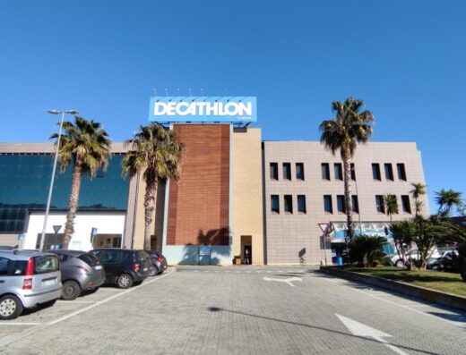 Vista esterna del negozio Decathlon Albenga con parcheggio auto, palme e cielo azzurro. Decathlon offre articoli sportivi, abbigliamento e accessori per moto e viaggi all'aperto.