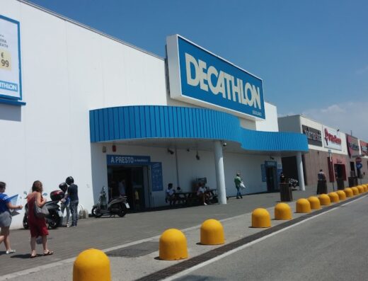 Decathlon Giugliano store front with customers entering, a scooter parked nearby, and yellow bollards lining the parking area.