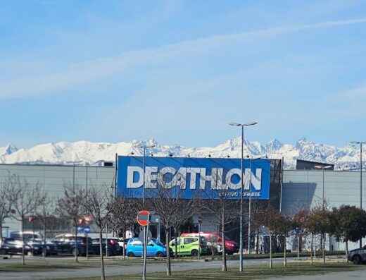 Decathlon Settimo Torinese store exterior with parking lot, visible signage and snowy mountains in the background on a sunny day.