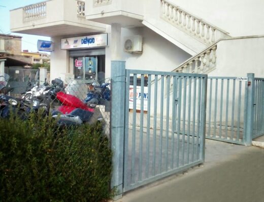 Dengio Racing officina riparazioni moto e scooter: vista esterna dell'officina con moto parcheggiate, ingresso con cancello in metallo e insegna ben visibile.