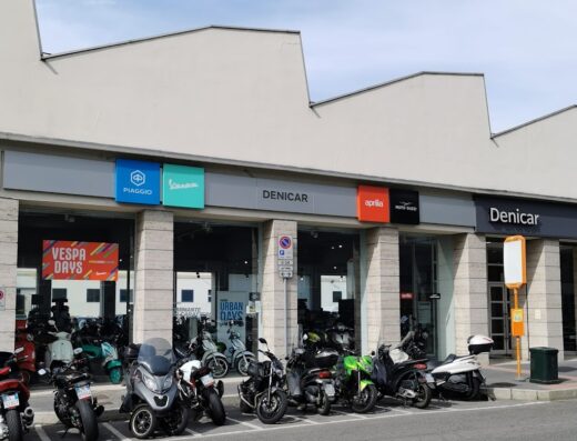 Concessionaria Denicar moto e scooter con esposizione di modelli Piaggio, Vespa, Aprilia e Moto Guzzi parcheggiati di fronte all'ingresso.