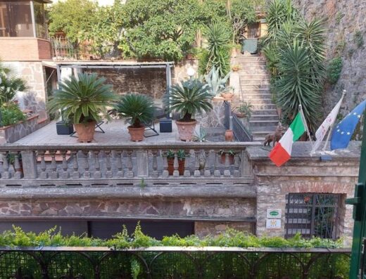 Vista dalla finestra sulla terrazza del Glam Resort, con piante in vaso, bandiere italiana ed europea, e scale in pietra.