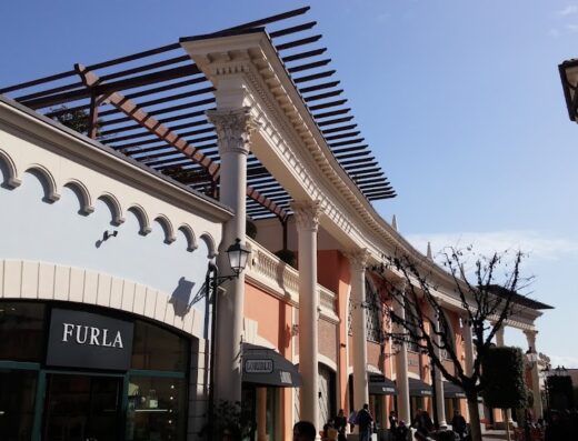 Veduta esterna del Designer Outlet Castel Romano con negozi di marchi come Furla e Michael Kors, architettura elegante e cielo azzurro.