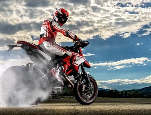Motociclista su Ducati Hypermotard rossa e bianca impenna con fumo dalla gomma, abbigliamento tecnico, casco rosso, cielo nuvoloso. Accessori e ricambi moto disponibili su Desmolupo.
