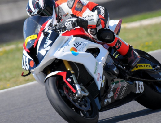 Pilota del Di Vora Andrea Racing Team in curva su moto da corsa con livrea bianca, rossa e nera, numero 133 ben visibile, sponsor tecnici Dunlop e HJC, abbigliamento tecnico e casco.