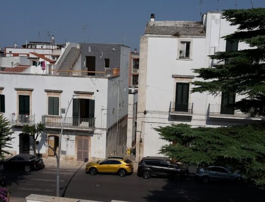 Vista di Dimora Intini, hotel di charme con architettura tradizionale pugliese, auto parcheggiate e vegetazione lussureggiante. Alberobello, Puglia.