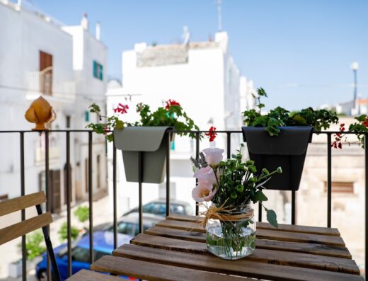 Balcone fiorito a Dimora Pozzo Epifani, con tavolo in legno, fiori in vaso e vista sulle case bianche di Puglia, ideale per un soggiorno rilassante e autentico.