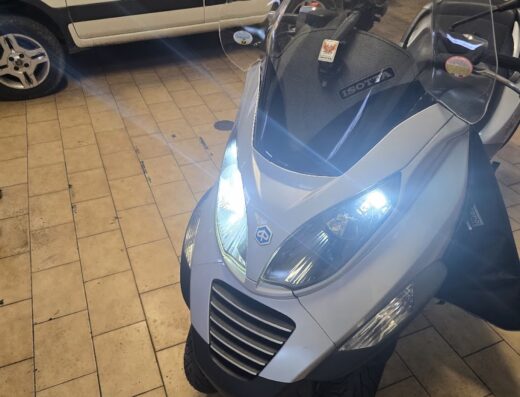Scooter Piaggio MP3 bianca con parabrezza Isotta, in un garage con auto sullo sfondo. Servizi e accessori moto disponibili.