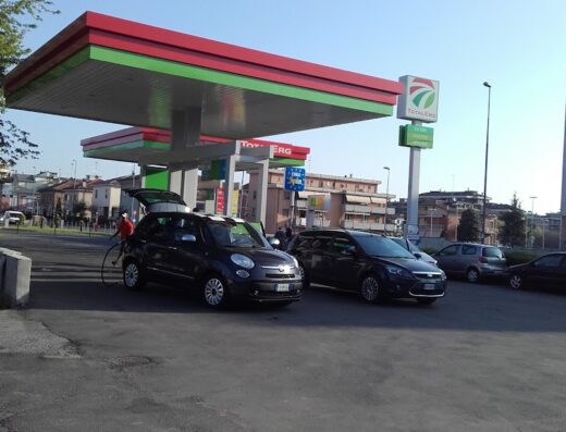 Distributore IP TotalErg con auto in rifornimento. Stazione di servizio con diverse auto parcheggiate. Zona urbana con edifici sullo sfondo.