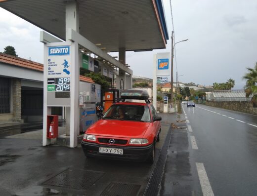 Auto rossa con portapacchi si rifornisce presso distributore IP, prezzi carburante visibili, viaggio in moto, accessori moto.